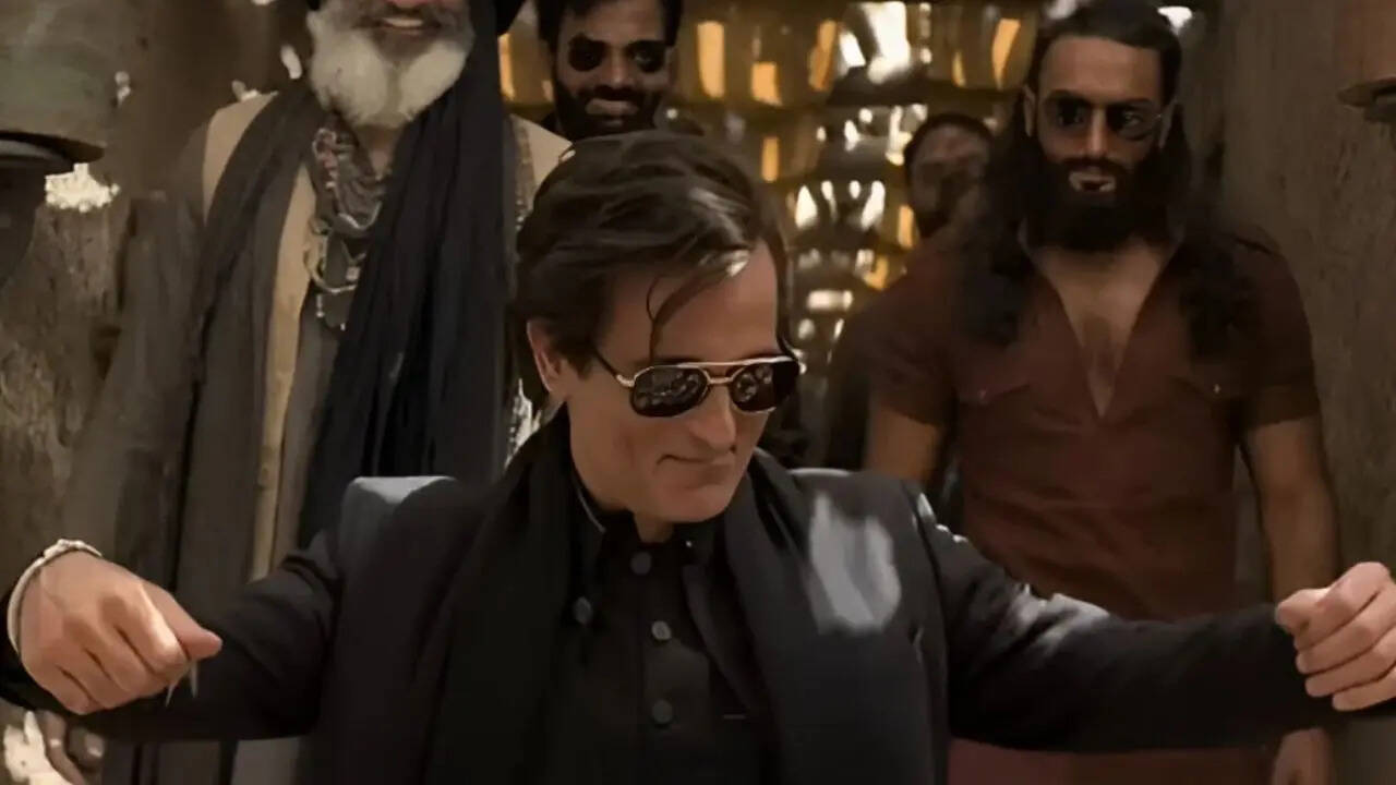 <strong>Dhurandhar Movie Viral Scene Pose Prompt 1</strong>- A confident, stylish man walking forward in a dramatic marketplace scene, wearing an all-black outfit with a long coat and sunglasses. Dust and debris fly around him, creating an action-movie effect. Behind him, two men walk in support — one in a brown kurta with long hair and sunglasses, the other in a dark jacket smiling. The background shows colorful market decorations, festive lanterns, and blurred buildings. Warm cinematic light.